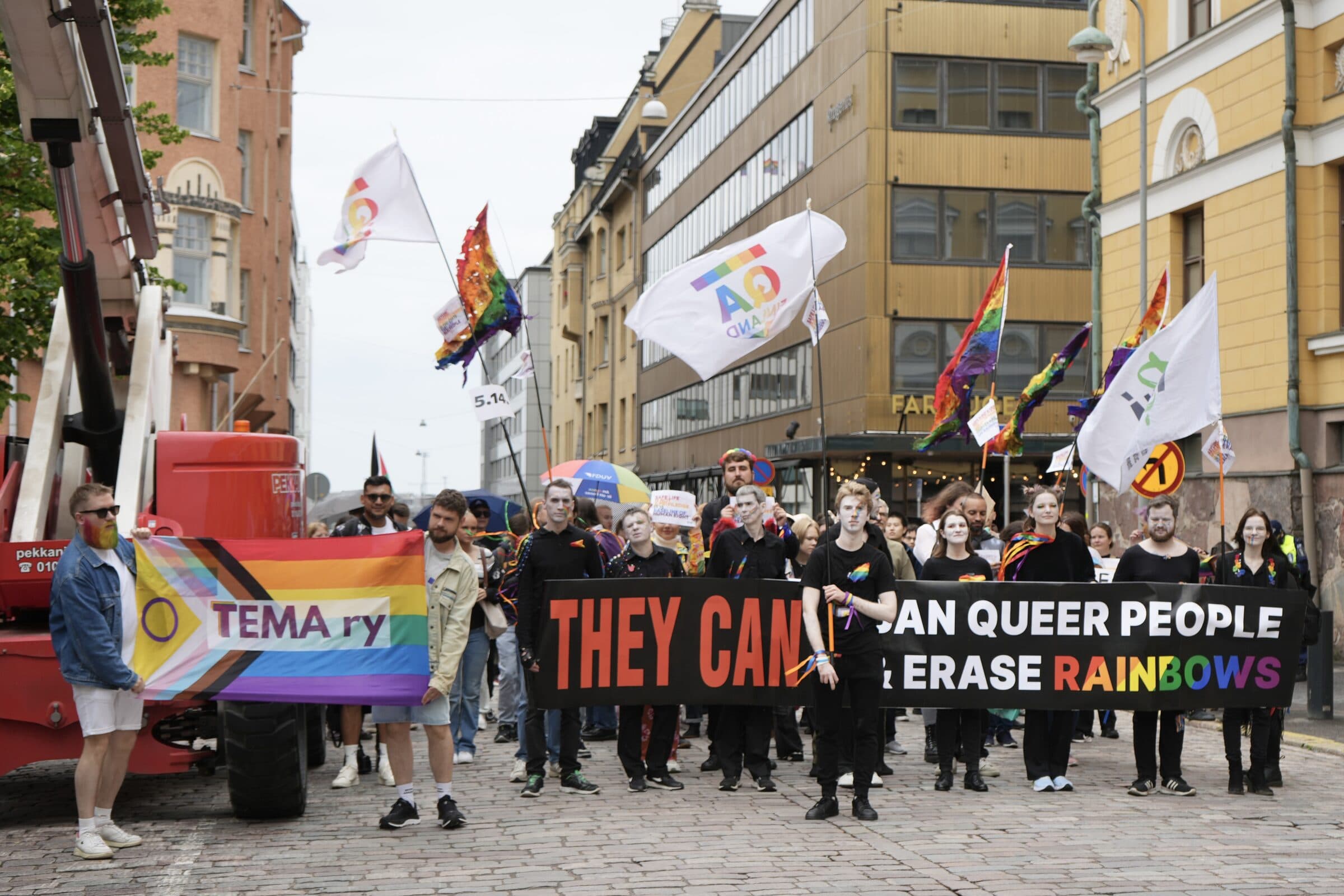 Tema ryn osallistujat kulkevat Pride-kulkueessa lippujen ja mielenosoituskyltin kanssa.